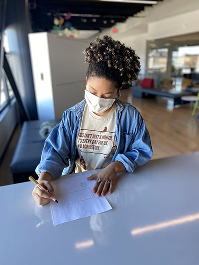 A young black woman wears a face mask while sketching.