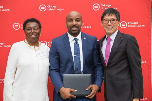 A black woman, black man and Asian man stand smiling in suits in front of a Chicago Urban League sign.