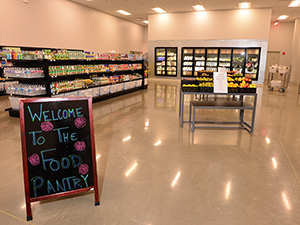 A brightly lit food pantry with well-organized and fully-stocked shelves with a sign that says “welcome to the food pantry” in colored chalk