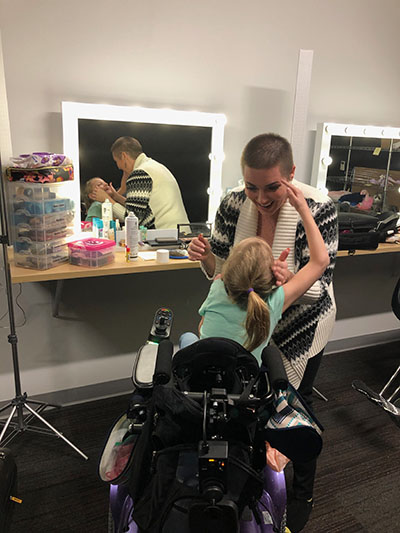 A makeup artist smiles as she applies makeup to the face of a young blonde girl in a wheelchair whose face can be seen smiling up at the artist in the mirror.