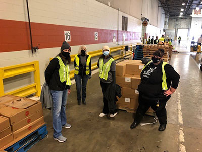Four Meijer team members wear reflective vests as they stand next to piles of boxes in a warehouse.