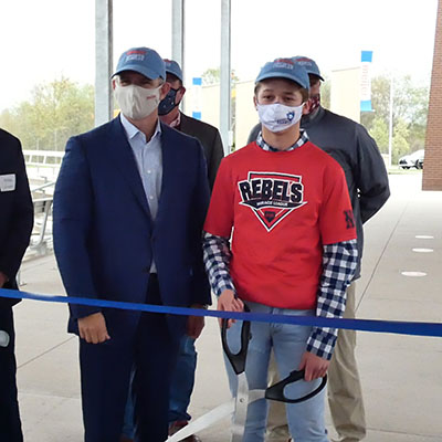 Rick Keyes, President and CEO of Meijer, stands next to a boy wearing a Miracle League t-shirt holding oversized scissors ready to cut the ribbon on the new Meijer Sports Complex.