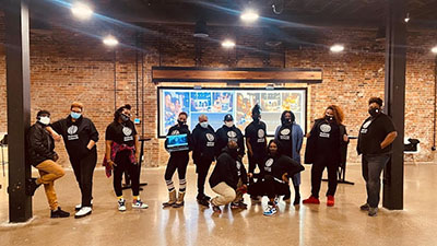 Eleven Black and African American BU Wellness Network employees stand in front of a projector screen posing in matching BU Wellness Network shirts.