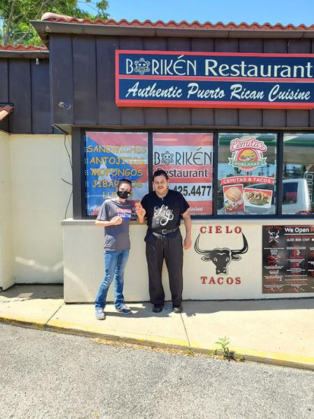 A Meijer team member wearing a T-shirt that reads “Stronger Together” stands next to a man wearing a hair net in front of a small restaurant with a sign that reads “Authentic Puerto Rican Cuisine”