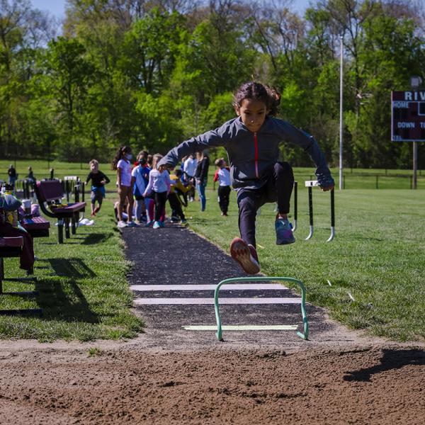 A girl jumps over a hurdle on a track while other children wait for their turn in the background
