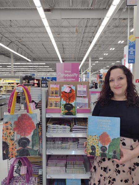 Misty Russian holding a notebook featuring her artwork next to a Women’s History Month display in a Meijer store.