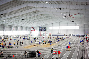 The spacious indoor track facility hosts athletes competing in a variety of track and field events including long jump, hurdles and pole vaulting while spectators watch from the stands.